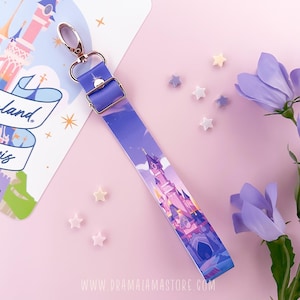 Puede incluir: Un cordón morado con un cierre y hebilla dorados, con una ilustración de un castillo. El cordón tiene una estética de cuento de hadas, con un fondo morado claro y adornos en forma de estrella. El sitio web www.dramalamastore.com es visible.