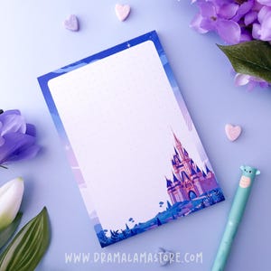Puede incluir: Un bloc de notas rectangular con un interior blanco de puntos y un borde azul que presenta una ilustración detallada de un castillo en la parte inferior. El bloc incluye el texto WWW.DRAMALAMASTORE.COM impreso en el centro inferior en blanco.