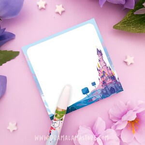 May include: A square sticky note with a white center and a border featuring a purple and blue castle illustration. A white pen with a colorful graphic design rests on the bottom edge of the note. The note includes the text WWW.DRAMALAMASTORE.COM.