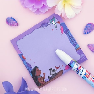 Peut inclure: Un bloc-notes carré violet illustré d'un personnage aux longs cheveux noirs et d'un raton laveur dans un canoë, avec un colibri au-dessus. Un stylo blanc avec des accents bleus et rouges repose sur le bloc-notes au centre violet clair.