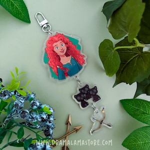 May include: An acrylic keychain featuring a character with long curly red hair in a blue dress, a secondary charm with three small black bear figures, and a third charm shaped like a bow and arrow, all connected by silver metal jump rings.