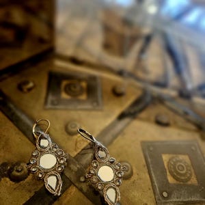 Könnte beinhalten: Ein Paar Ohrhänger mit einer geometrischen Anordnung aus kleinen kreisförmigen und tropfenförmigen Spiegeln, die von Metallfäden umrandet sind. Jeder Ohrring hängt an einem goldfarbenen Metallhaken und hat einen runden Spiegel in der Mitte.