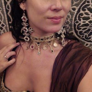 Puede incluir: Un collar tipo gargantilla dorado con detalles colgantes circulares y en forma de diamante con piedras transparentes y rojas, junto con pendientes largos a juego y un top negro con ribete de malla metálica dorada.
