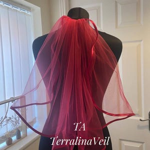 May include: A red bridal veil on a black mannequin. The veil is made of sheer fabric with a satin trim. The text "TA TerralinaVeil" is visible on the lower right. A wedding accessory, the veil is a vibrant red.