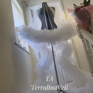 Op de afbeelding: Een witte bruidssluier met een gelaagd, gerimpeld ontwerp wordt op een mannequin getoond. De sluier heeft een lange sleep en een volledig, pluizig rokachtig detail. De tekst "TA TerralinaVeil" is zichtbaar.