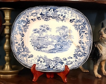May include: An oval ceramic platter featuring a blue and white transferware pattern. The centre depicts a landscape with a bridge, trees, and buildings, while the border is decorated with intricate floral motifs and a dotted edge design.