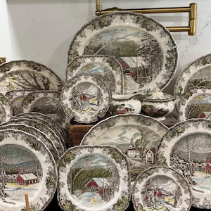 May include: A collection of ceramic dinnerware featuring winter landscape scenes with red buildings and snow-covered trees. Each piece has a decorative border of green leaves and brown vines. The set includes various plates, a gravy boat, and a lidded bowl.