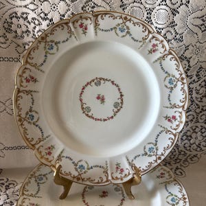 May include: White porcelain plates with a scalloped edge and gold trim. The plates feature a floral design with pink and blue flowers. The plates are displayed on a gold stand, showcasing their elegant design.