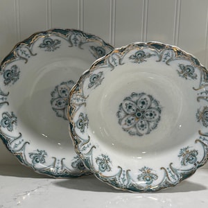 May include: Two white ceramic bowls featuring a scalloped edge and a decorative pattern in shades of dark teal, grey, and gold. The center of each bowl displays a symmetrical floral motif, while the rims are adorned with intricate scrollwork.