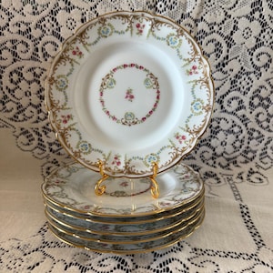 May include: A stack of white porcelain plates with a floral design and gold trim. The plates have a scalloped edge and are displayed on a gold plate stand. The floral pattern includes pink, blue, and green accents.