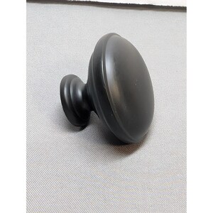 May include: A matte black cabinet knob featuring a smooth, rounded, mushroom-shaped head and a tapered, circular base. The hardware has a solid, uniform dark finish with a clean, curved profile designed for attachment to furniture or cabinetry.
