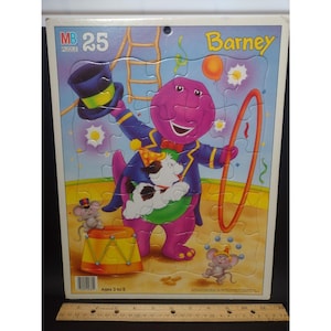 Puede incluir: Un rompecabezas de 25 piezas de Barney el dinosaurio. El dinosaurio morado está vestido de mago, sosteniendo un sombrero de copa y un aro. El rompecabezas presenta un perro, ratones, un tambor y el texto "Ages 3 to 5".