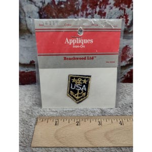 May include: A small shield-shaped iron-on applique patch featuring a black background with gold trim, a gold anchor, three gold stars, and the white text USA. The patch is inside a clear plastic package labeled Appliques Iron-On Beachwood Ltd.