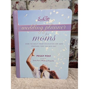 May include: A light blue hardcover book titled Emily Post's Wedding Planner for Moms: How to Help Your Daughter or Son Prepare for the Big Day by Peggy Post. The cover features a person in a white top reaching up amidst falling confetti.