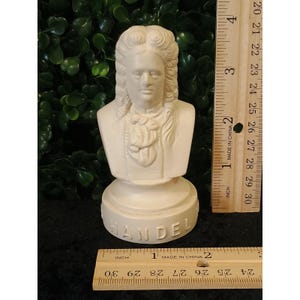 May include: A white matte bust of the composer Handel, featuring a detailed wig and cravat. The base is inscribed with the name HANDEL. A wooden ruler is positioned vertically and horizontally, showing the bust stands approximately 4.5 inches tall.