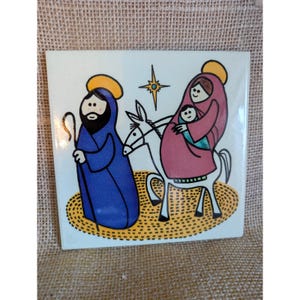 May include: A square ceramic tile featuring a colorful illustration of a nativity scene. The design shows a figure in a blue robe with a staff, a donkey carrying a figure in a pink robe holding a baby, and a yellow star above, all on a yellow ground.