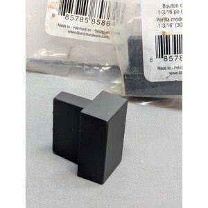 May include: A matte black rectangular cabinet knob with a clean, geometric design. The knob features a solid, block-like shape and is positioned in front of its original clear plastic packaging, which displays a barcode and the text 1-3/16 inches.