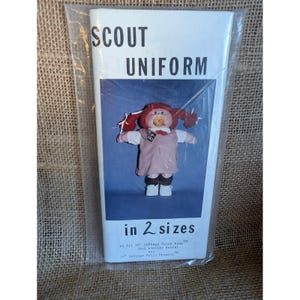May include: A Scout Uniform sewing pattern package in a clear plastic sleeve. The white packaging features the text SCOUT UNIFORM and in 2 sizes, with an image of a doll wearing a tan dress, white shirt, and white shoes. Text notes it fits 16 inch dolls.