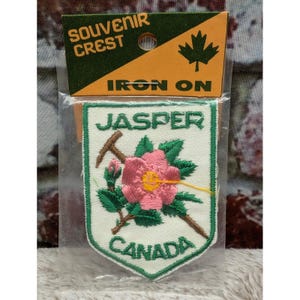May include: An iron-on souvenir crest patch featuring the text Jasper Canada. The shield-shaped patch has a white background with a green border, a pink flower with green leaves, and a brown branch. It is packaged in a clear plastic sleeve.