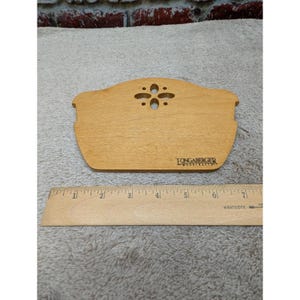 May include: A light brown wooden basket lid with a cutout floral design and the text Longaberger Woodcrafts printed in black. A wooden ruler is placed in front, showing the item is approximately 19 cm wide at its widest point.