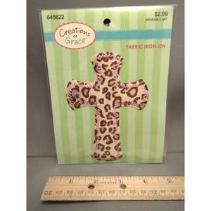 May include: A fabric iron-on cross with a leopard print design. The cross is tan with brown spots and pink trim. The packaging includes the text "Creations of Grace" and "Wearable Art". A ruler is at the bottom of the image.