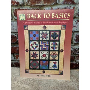 May include: A softcover book titled Back to Basics: A Quilter's Guide to Patchwork and Applique by Donna Wilder. The cover features a maroon border and a central grid displaying twelve distinct quilt block patterns in various colors and designs.