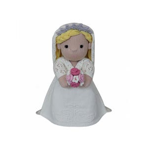 May include: A knitted bride doll, wearing a white dress and veil, with blonde hair and a floral crown. The doll holds a small bouquet of pink and white flowers. The doll's face has embroidered features, and the dress has a detailed pattern.