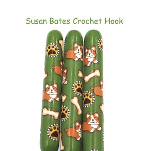 May include: Three green crochet hooks with a pattern of brown and white corgis and bone designs. The text "Susan Bates Crochet Hook" is written above the hooks.