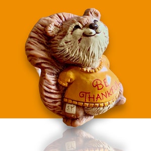 May include: A brown and tan squirrel figurine wearing a yellow shirt with the words "Be Thankful" in red lettering. The squirrel is smiling and has a heart shape on its shirt.