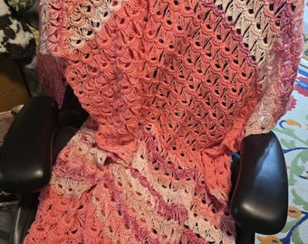May include: A handmade crochet blanket featuring an intricate shell stitch pattern in a gradient of colors ranging from soft cream and pale pink to vibrant coral and deep magenta, creating a textured, openwork design that drapes over a chair.