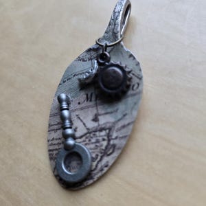 May include: A handcrafted pendant made from a repurposed spoon, featuring a map design and small metal charms. The pendant includes a magnifying glass charm and a sunburst charm, with a vintage aesthetic.
