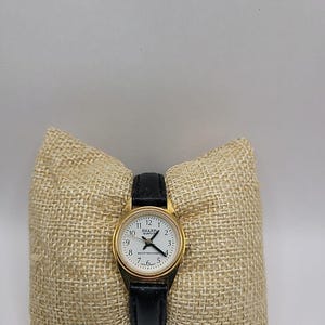 May include: A gold-toned Sharp quartz watch with a white face and black hands and numbers. The watch has a black leather band and is displayed on a beige burlap pillow. The watch face reads "SHARP QUARTZ" and "WATER RESISTANT".
