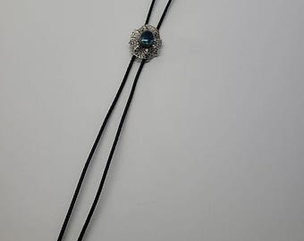 May include: A bolo tie featuring a black braided cord with silver-toned metal tips. The central slide is a circular, intricate silver-toned filigree setting holding a single, oval-shaped, faceted blue gemstone in the center.