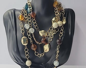 May include: A multi-strand necklace with gold-tone chains and a variety of beads. The necklace features clear, amber, brown, and blue beads, along with gold-colored, irregularly shaped pendants. The necklace is displayed on a black bust.