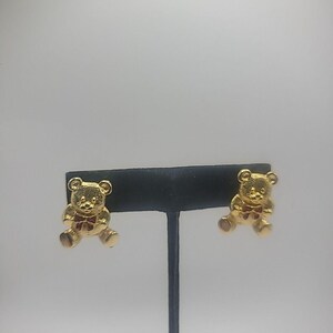 May include: Gold-toned teddy bear earrings. Each earring features a detailed bear design with a red bow tie. The earrings are displayed on a black jewelry stand against a white background.