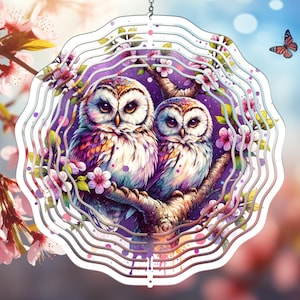 May include: A colorful metal wind spinner with two owls perched on a branch with pink flowers. The background is a purple and white watercolor design.