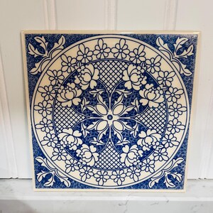 May include: Square ceramic tile with a blue and white floral design. The tile features a central floral medallion surrounded by a ring of flowers and a lattice pattern. The corners have decorative floral accents.