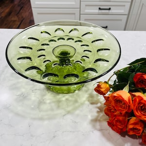 May include: A green glass cake stand with a pedestal base. The stand has a circular top with decorative cutouts and a central raised area. The stand is approximately 10 inches in diameter. A bouquet of orange roses is in the background.