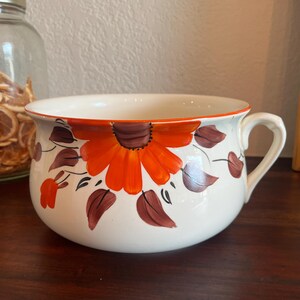 May include: White ceramic bowl with a large orange and brown floral design. The bowl has a handle and a decorative orange rim. The floral design includes orange petals and brown leaves. The bowl is likely for serving or display.