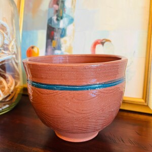 Op de afbeelding: Een handgemaakte, roodbruine keramische kom met een blauwgroene streep. De kom heeft een textuuroppervlak met golfachtige patronen. De kom is rond met een brede opening en een licht uitlopende rand. Hij wordt waarschijnlijk gebruikt voor het serveren van voedsel.