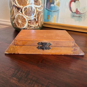 May include: A small, rectangular wooden box with a hinged lid and a decorative metal clasp. The box is light brown and sits on a slightly larger base. The background includes a glass jar filled with dried orange slices and a framed painting.