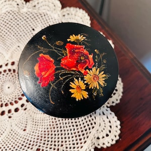 May include: A round, black decorative box with a painted floral design. The flowers are red poppies and yellow daisies with green leaves. The box is sitting on a white lace doily.