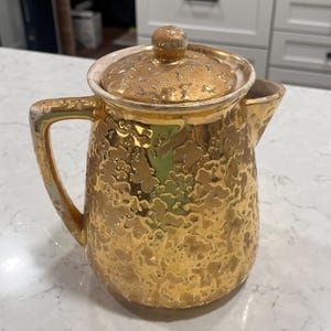 Op de afbeelding: Een vintage goudkleurige keramische koffiepot met een textuuroppervlak en een bijpassende deksel. De pot heeft een handvat en een tuit. De deksel is bekroond met een kleine, ronde knop. De koffiepot is waarschijnlijk uit het midden van de eeuw.