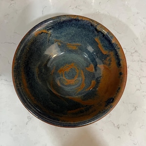 May include: A handmade ceramic bowl with a dark blue interior and a reddish-brown exterior. The bowl has a glossy finish and a unique, organic design. The interior features swirling patterns of orange and blue.