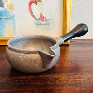 May include: A small, gray metal saucepan with a spout and a black handle. The handle is curved and attached to the side of the pan. The pan is sitting on a dark wooden surface.