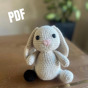 May include: A crochet bunny toy with light beige yarn, long floppy ears, black plastic eyes, and a stitched pink nose. The bunny has a small black spherical tail visible on the side. The text PDF is displayed in the upper left corner.