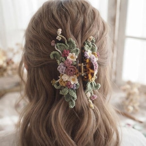 May include: A tortoise shell hair claw clip adorned with intricate crochet flowers and leaves in shades of sage green, dusty rose, cream, and lavender. The floral arrangement is attached to both sides of the clip, featuring small blossoms.
