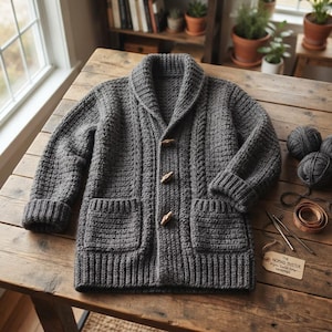 May include: A charcoal gray knit cardigan featuring a shawl collar, long sleeves with ribbed cuffs, two front patch pockets, and three wooden toggle buttons. A tag labeled The Nomad Duster is placed nearby with yarn balls and a crochet hook.