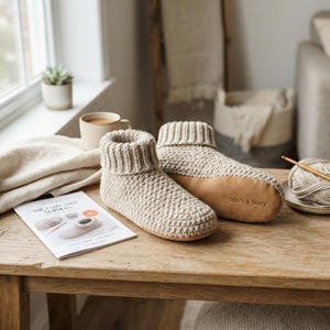Könnte beinhalten: Ein Paar beigefarbene Strickhausschuhe mit gerippten Bündchen und hellbraunen Wildledersohlen, auf denen der Schriftzug Stitch & Story zu sehen ist. Die Hausschuhe liegen neben einer Anleitung mit dem Titel The Cozy Step Slippers und Garn.