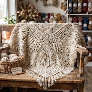 May include: A beige crochet shawl featuring a detailed eagle motif with spread wings and a fringed border. The shawl is draped over a wooden chair next to a woven basket containing yarn balls and a tag labeled Eagle Shawl Crochet Pattern.